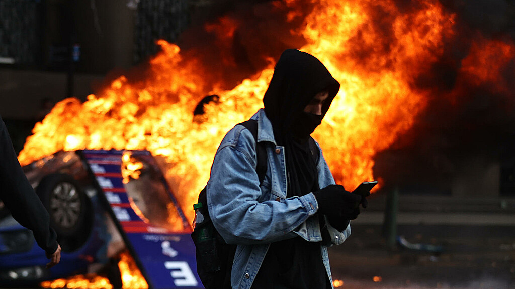 Una persona observa su celular mientras se da un incendio a sus espaldas durante enfrentamientos entre la policía