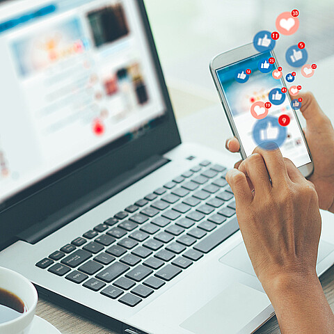 Computador en el fondo y una persona navegando en su celular en las redes sociales