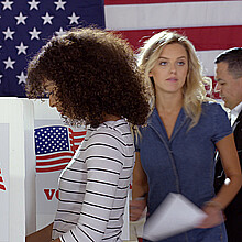 Four people of different demographics, young Hispanic woman in front, filling in ballots and casting votes in booths at polling station